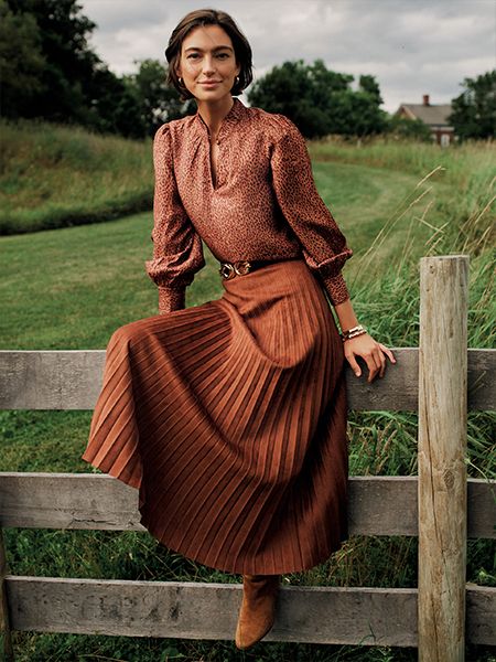 A woman with short dark hair smiles while sitting on a wooden fence, wearing a brown animal print blouse, a pleated rust midi skirt, and brown boots.