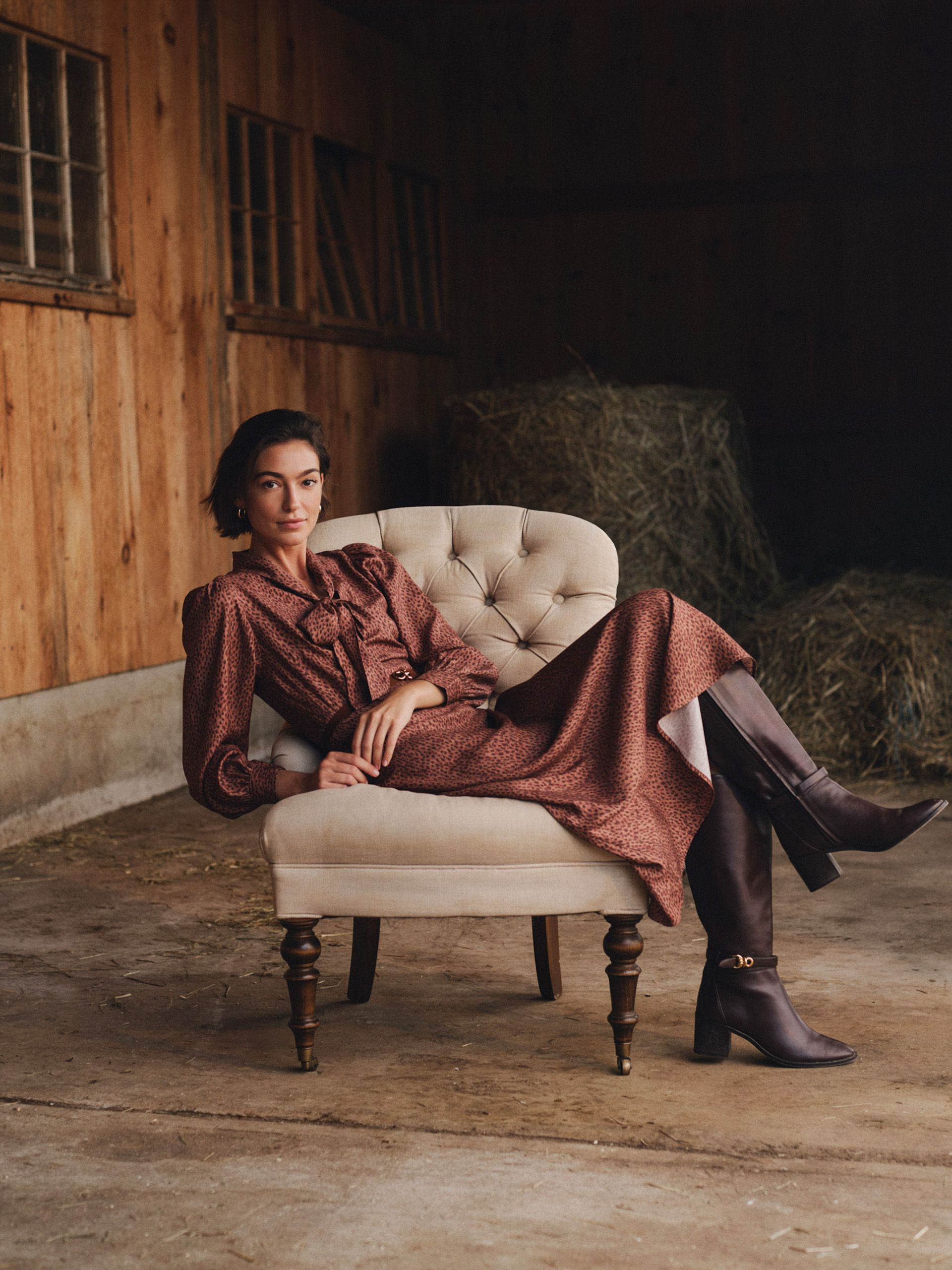 A woman in a rust-colored dress and tall dark boots poses in a cream armchair inside a barn with hay bales.