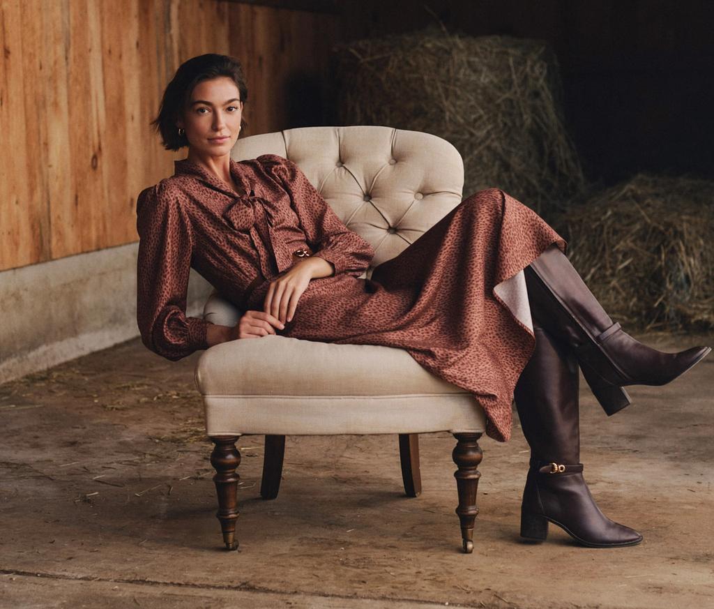 A woman in a rust-colored dress and tall dark boots poses in a cream armchair inside a barn with hay bales.