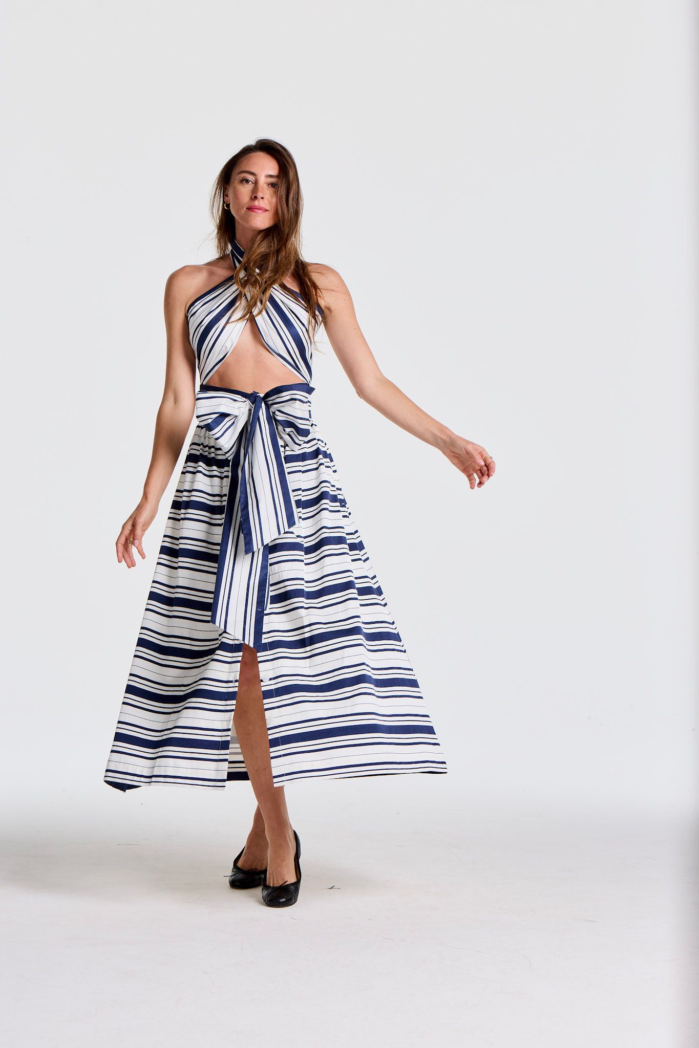 A woman wears a blue and white striped halter top and matching long skirt with a bow and slit, posing on a white background.