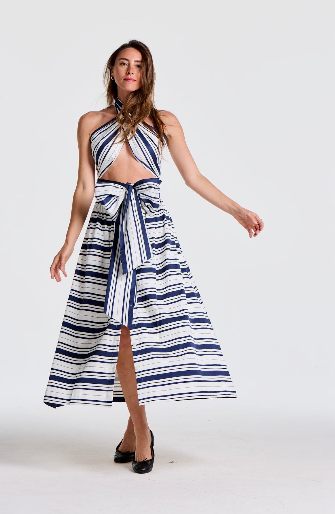 A woman wears a blue and white striped halter top and matching long skirt with a bow and slit, posing on a white background.