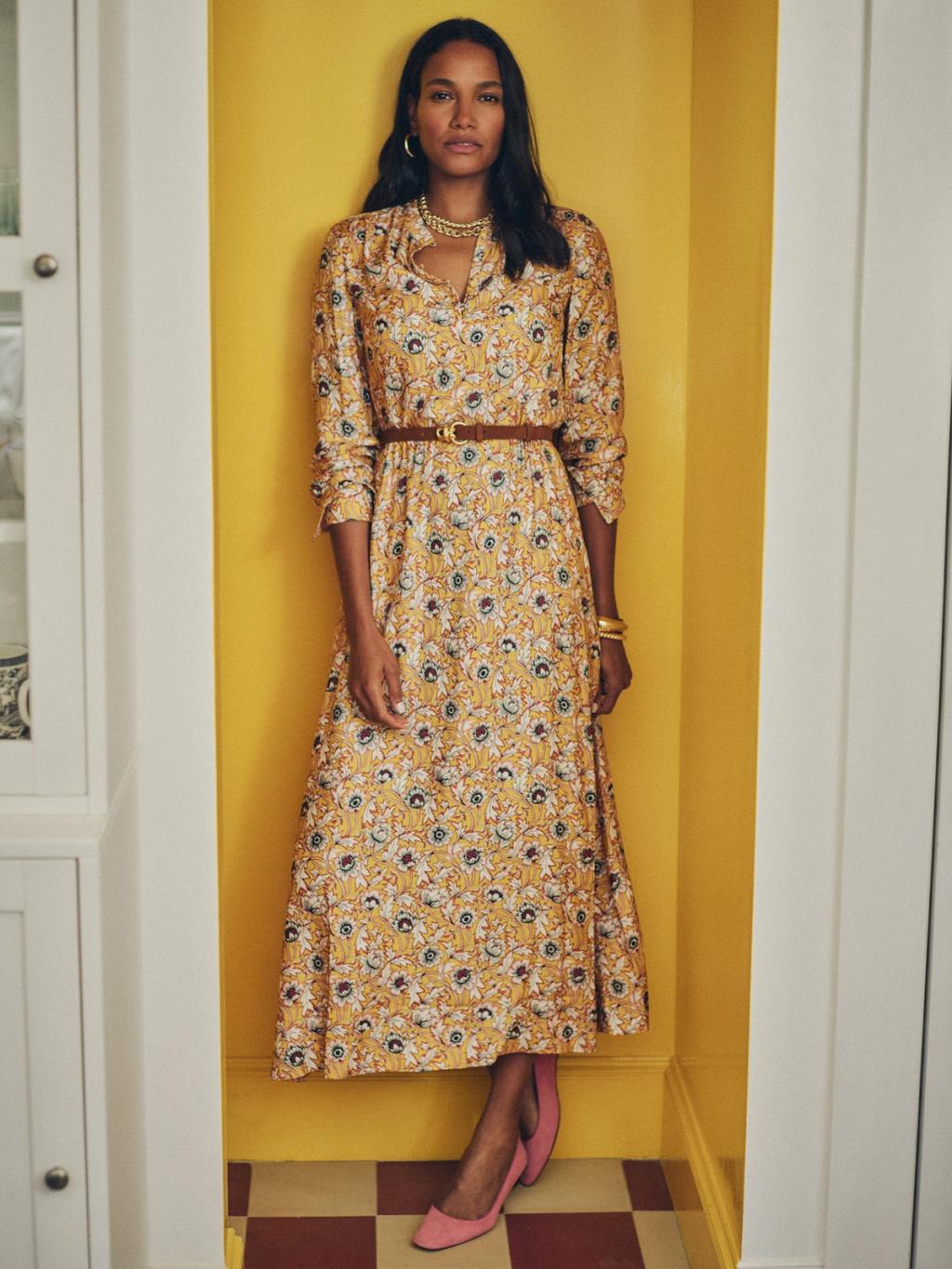 A woman stands in a yellow nook, wearing a yellow floral midi dress with a brown belt and pink pointed-toe flats.