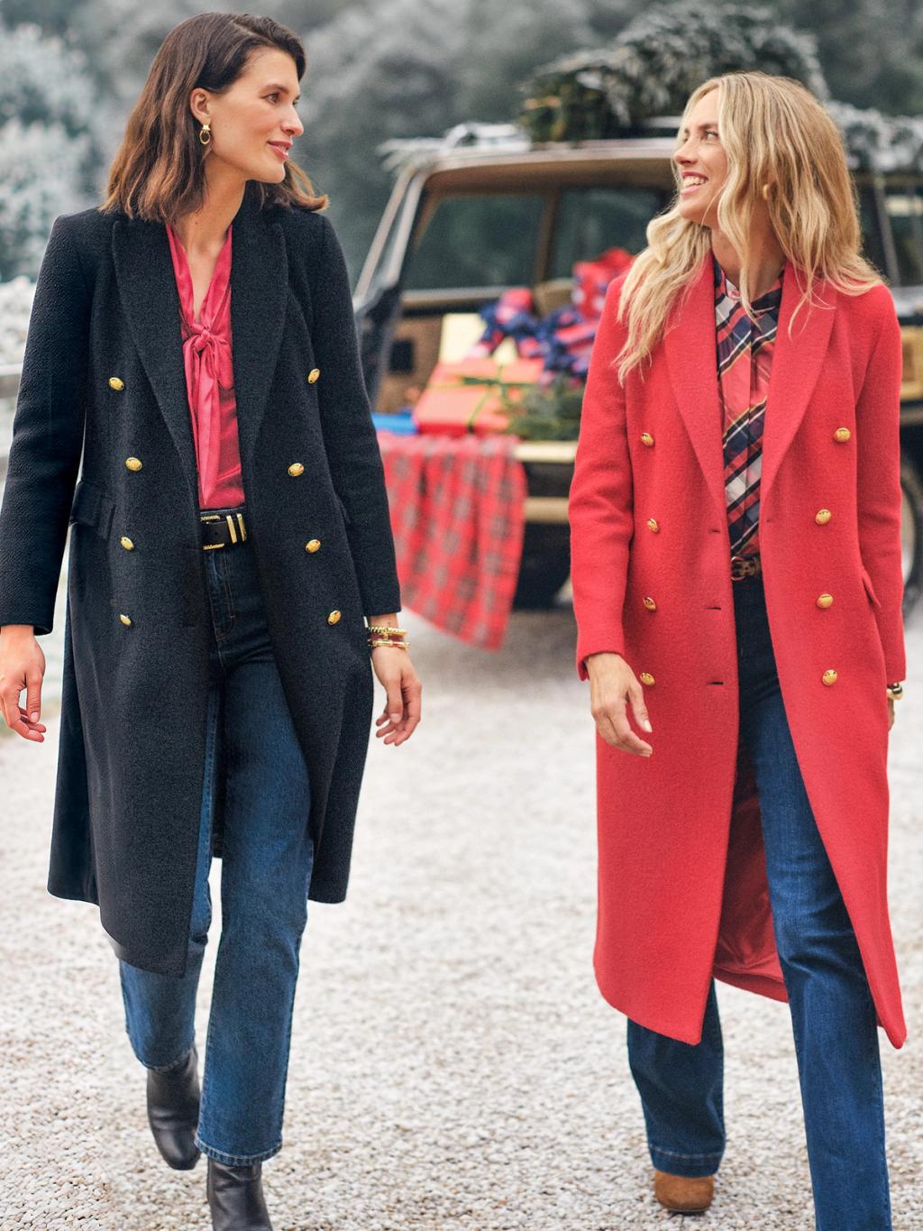 Two smiling women in dark gray and red double-breasted coats walk past an SUV with a Christmas tree and gifts.