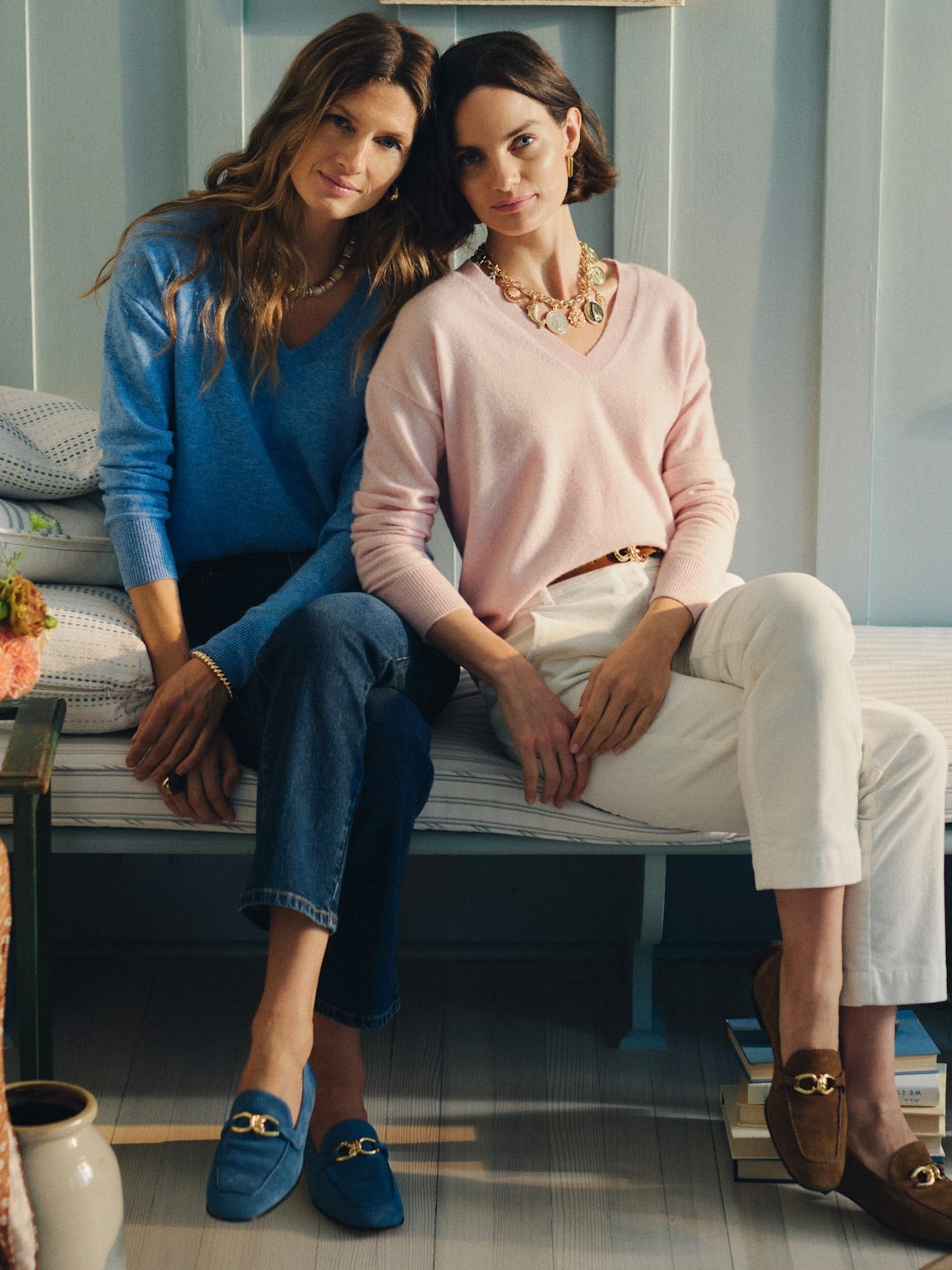 Two women in sweaters and loafers sit on a striped bench; one wears blue, the other wears pink and a chunky gold necklace.