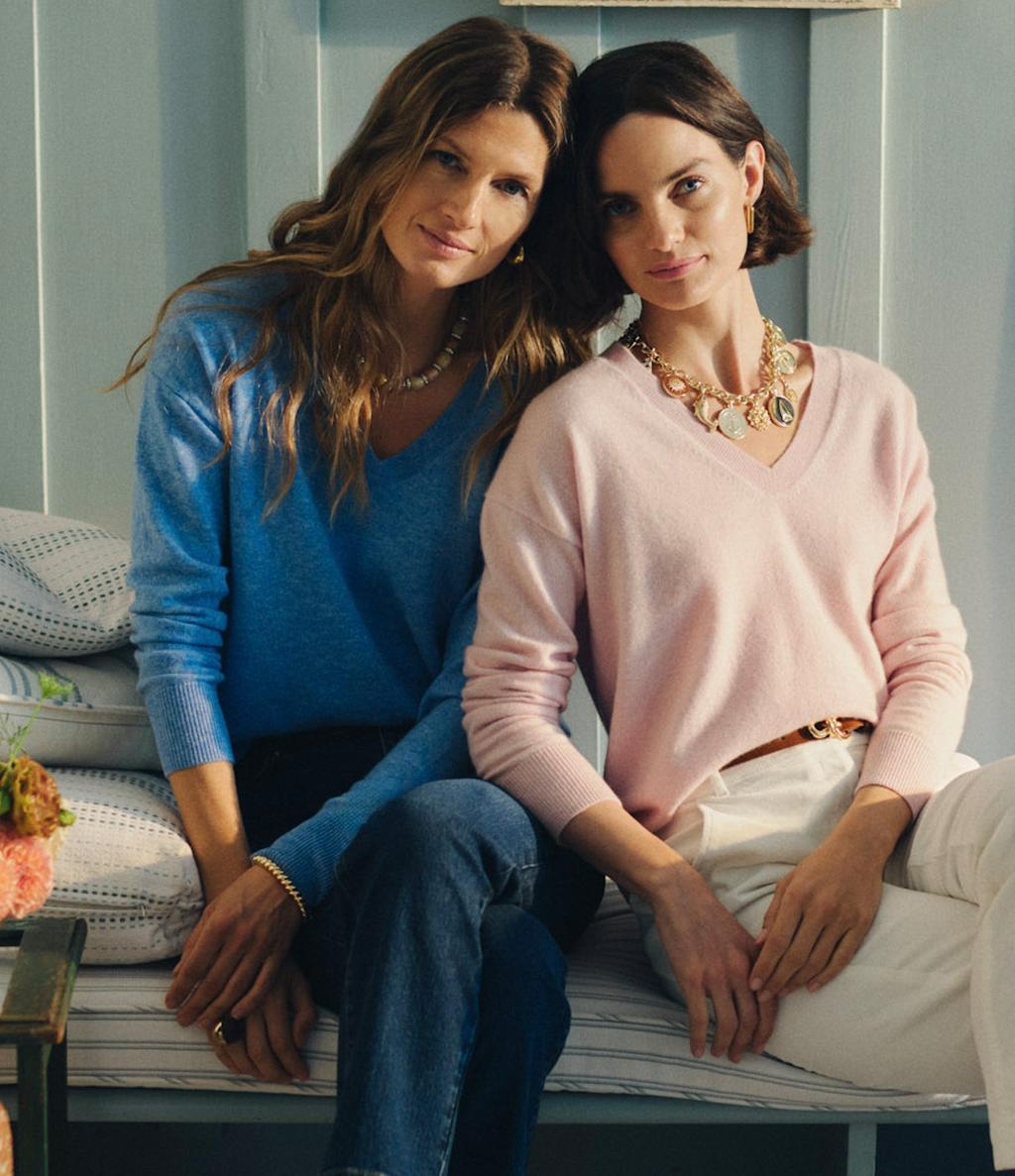Two women in sweaters and loafers sit on a striped bench; one wears blue, the other wears pink and a chunky gold necklace.