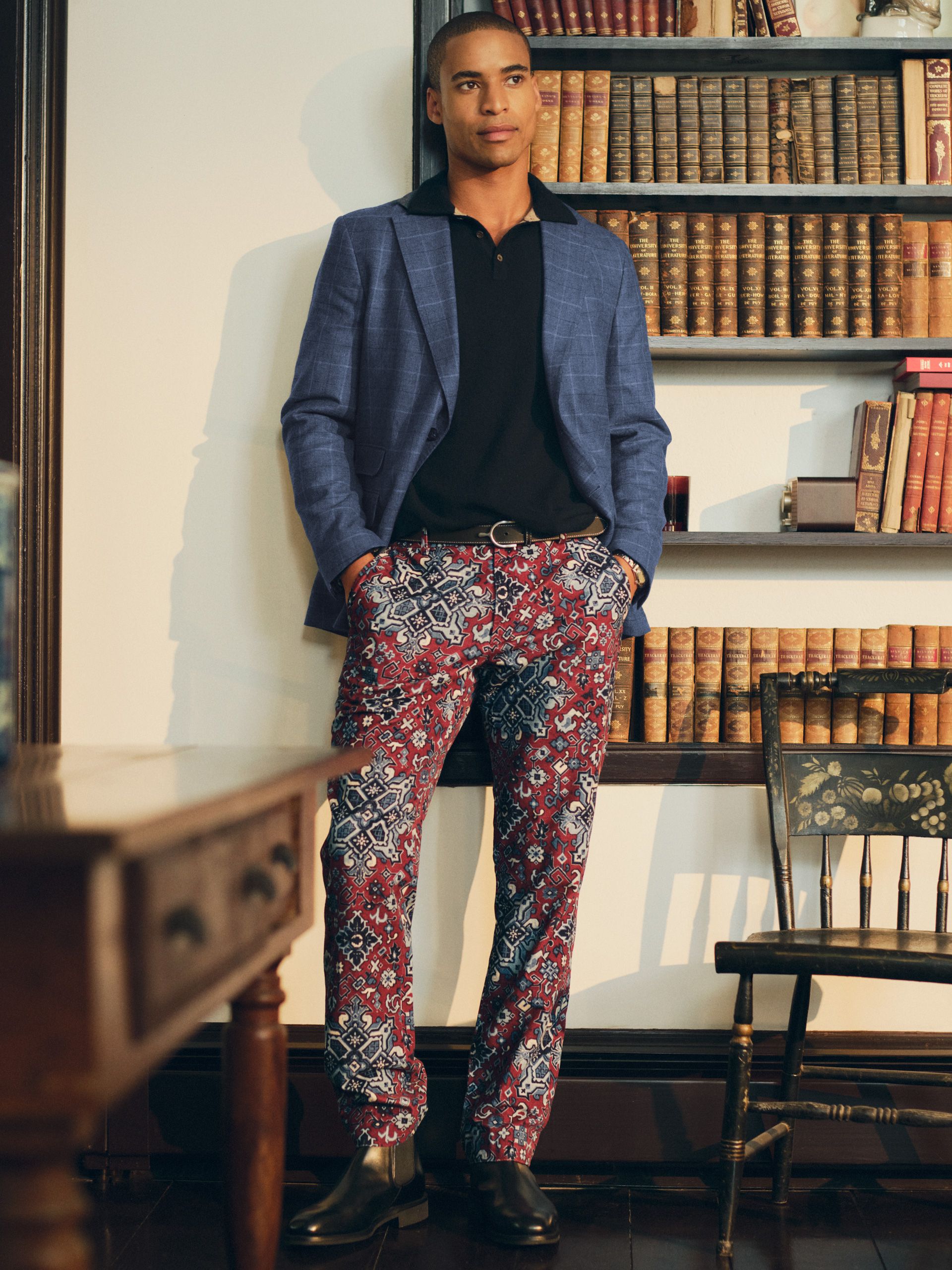 A man in a blue plaid blazer, black polo, and red paisley patterned pants stands in a library.