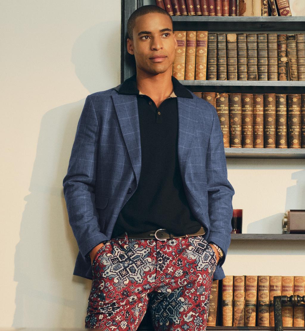 A man in a blue plaid blazer, black polo, and red paisley patterned pants stands in a library.