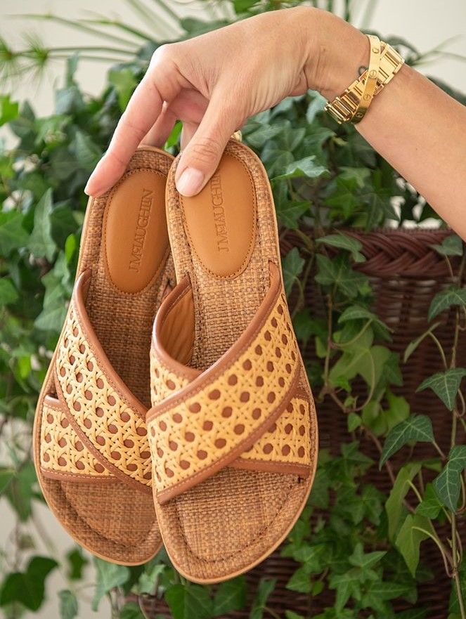 woman's hand holding out a pair of perforated leather and rattan cross strap slide sandals