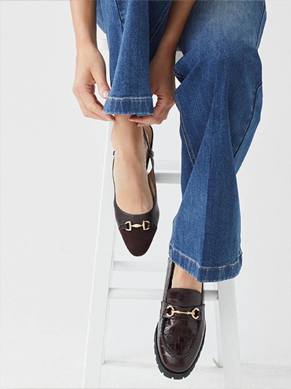 Two legs sitting on a chair wearing denim with a black loafer on one foot and a black ballet flat on the other.