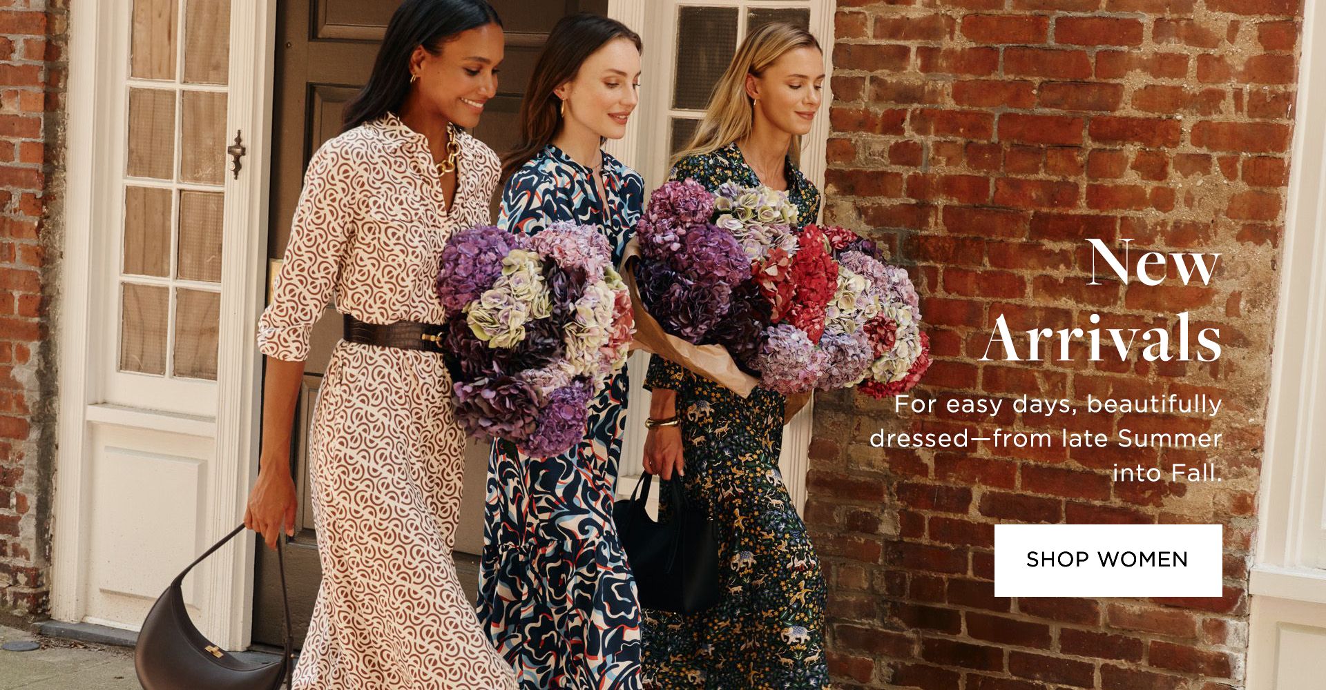 Three women standing together outdoors holding large bouquets of colorful flowers including purple, pink, red, and white blooms. They're wearing patterned dresses - one in a geometric print, one in an abstract blue and pink pattern, and one in a dark floral print. They're positioned in front of a brick building with white doors and windows. Text overlay reads "New Arrivals" and "For easy days, beautifully dressed—from late Summer into Fall" with a "SHOP WOMEN" button.