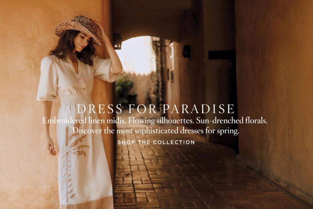 woman standing against tan wall wearing tan linen dress with embroidery detail and straw hat