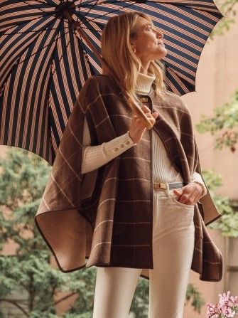 woman standing outside wearing cream sweater and pants, a brown windowpane cape and holding an umbrella