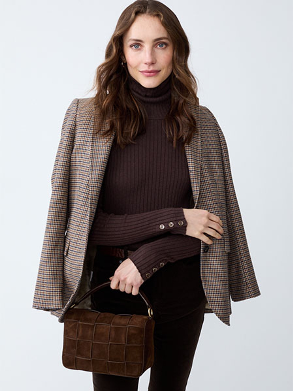 Women with brown hair holding a brown leather handbag while wearing a brown turtleneck and a patterned blazer draped over shoulders
