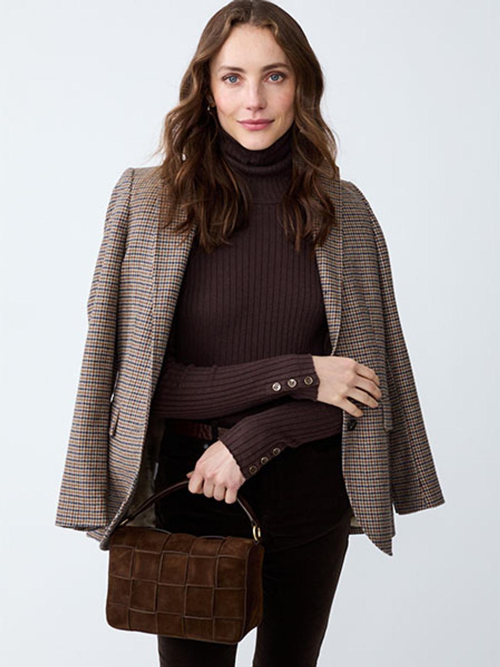 Women with brown hair holding a brown leather handbag while wearing a brown turtleneck and a patterned blazer draped over shoulders