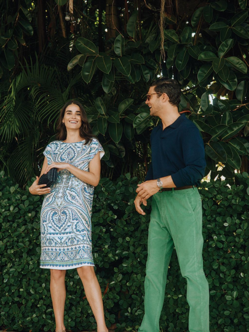 Female model in printed dress with wicker clutch. Male model in navy sweater and green pants.
