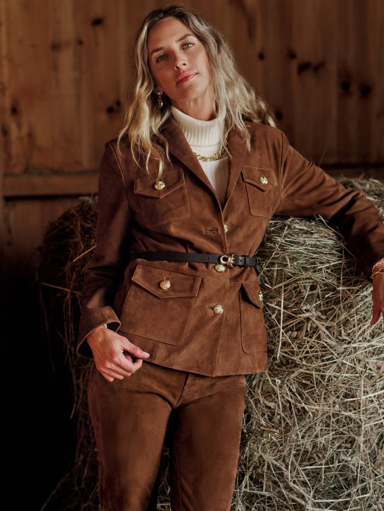 A woman in a brown suede jacket with gold buttons and matching pants, a white turtleneck, and a gold chain, leans on a hay bale in a barn.
