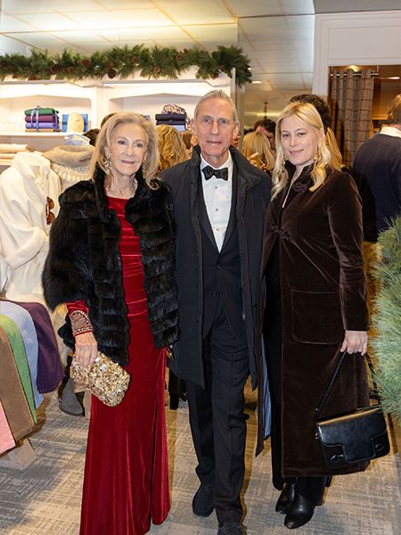 Three people in formal attire pose together at an upscale holiday event or party. Holiday decorations including garland are visible in the background of what looks like a retail or boutique setting.