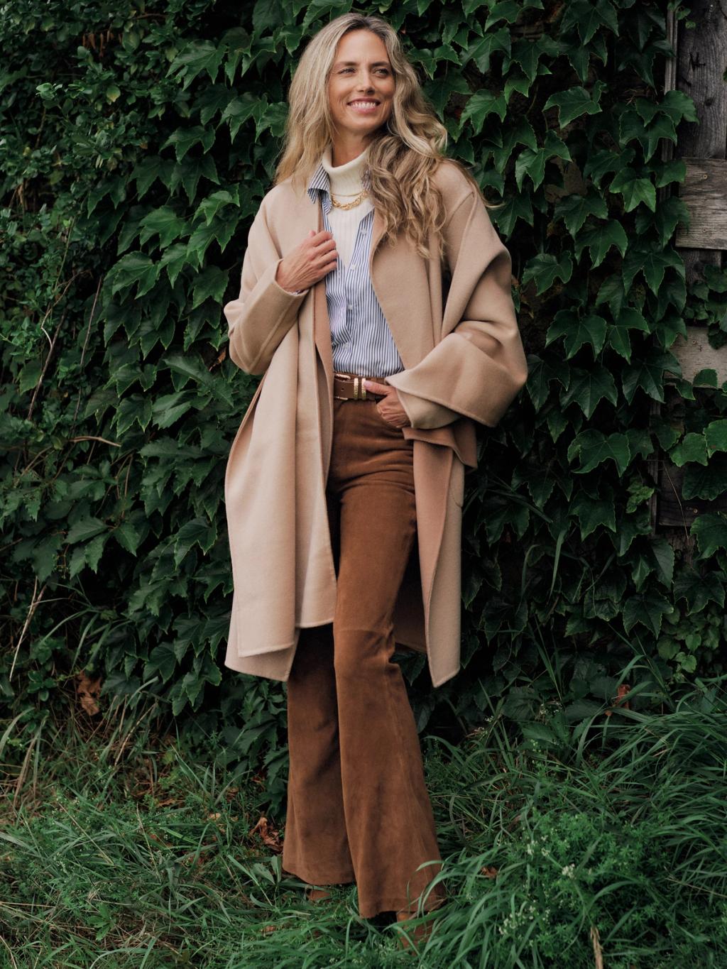 A smiling woman in a camel coat, striped shirt, and brown suede pants stands against a green ivy wall.
