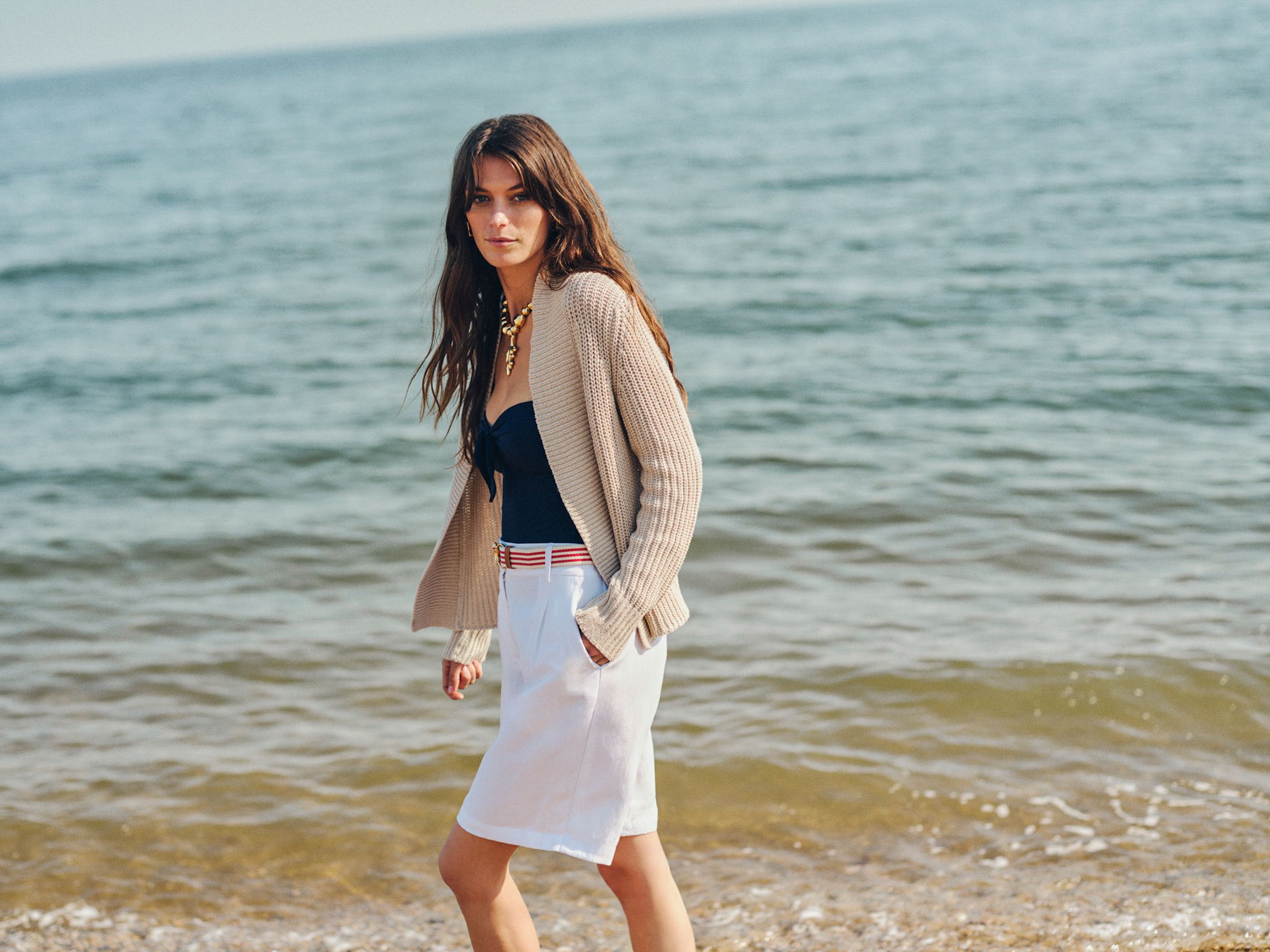 A woman walks on a beach wearing a beige cardigan, navy top, and white shorts.