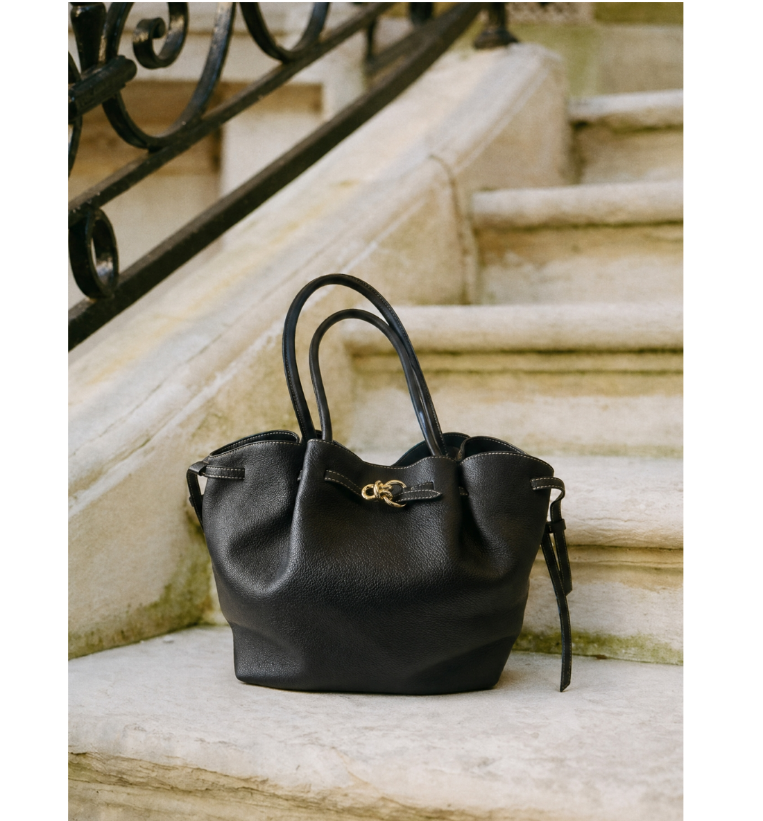 Black leather handbag with a gold clasp and top handles on stone steps.