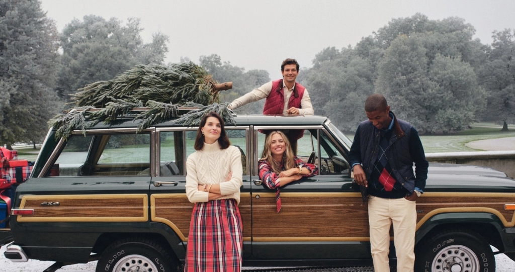 Four people pose with a Christmas tree strapped to a vintage woody wagon in a snowy landscape.