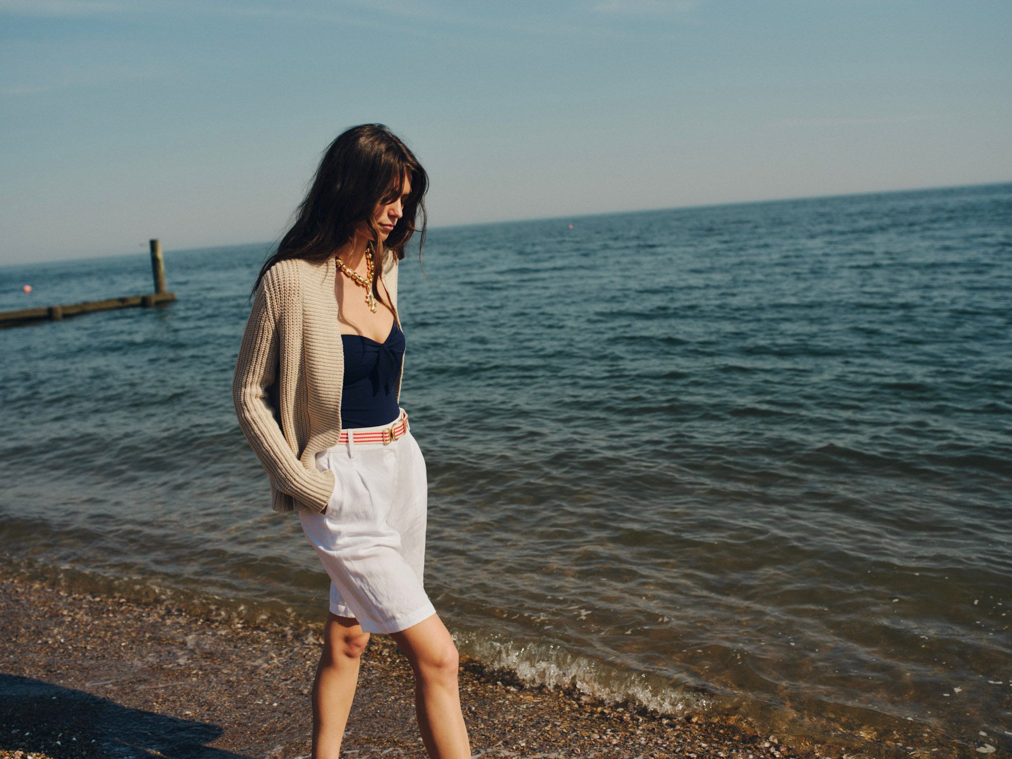 A woman in a beige cardigan, navy top, and white shorts walks along a beach with the ocean and a pier in the background.