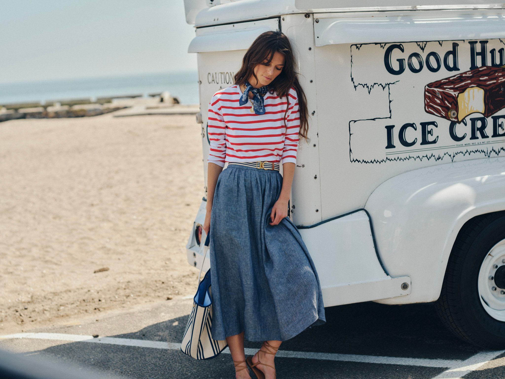 A woman in a red and white striped shirt and blue skirt leans against a white ice cream truck on a sandy beach.