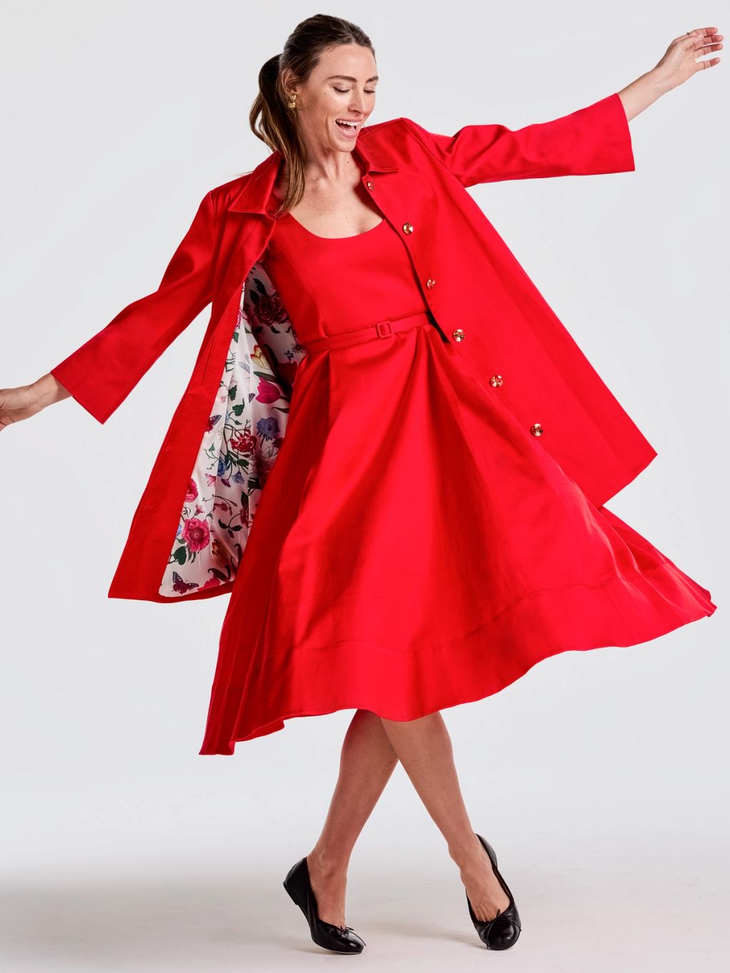 A smiling woman in a red dress and an open red coat with a floral lining, striking a joyful pose.