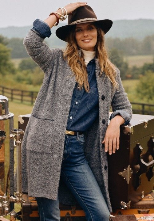 woman standing outside leaning on steamer trunk wearing jeans, a denim shirt, a brown cowboy hat and a gray knit cardigan coat layered over