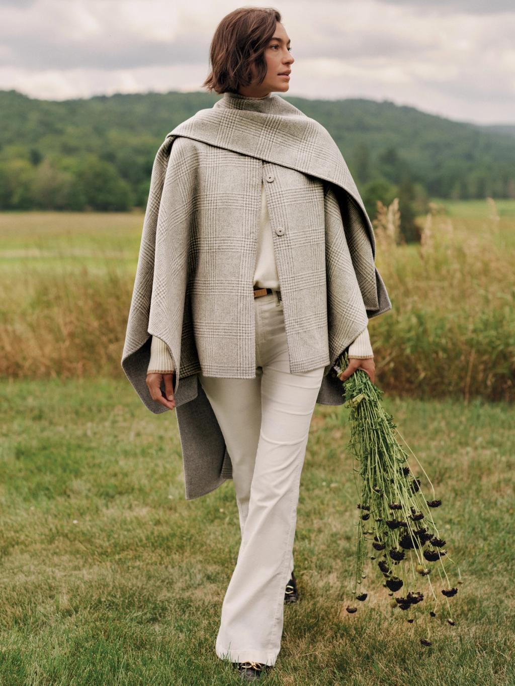 A woman in a gray plaid cape and light pants walks through a grassy field, holding dark flowers.