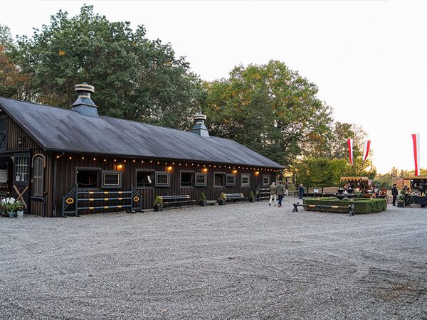 A rustic dark wooden barn-style building with a long slanted metal roof and two cupolas sits in a gravel courtyard during early evening. String lights are hung along the building's facade, which features multiple windows. Large trees with green and autumn foliage frame the scene in the background. People can be seen gathering in the outdoor space, with what appears to be additional seating areas and tall decorative banners or flags visible on the right side of the courtyard.