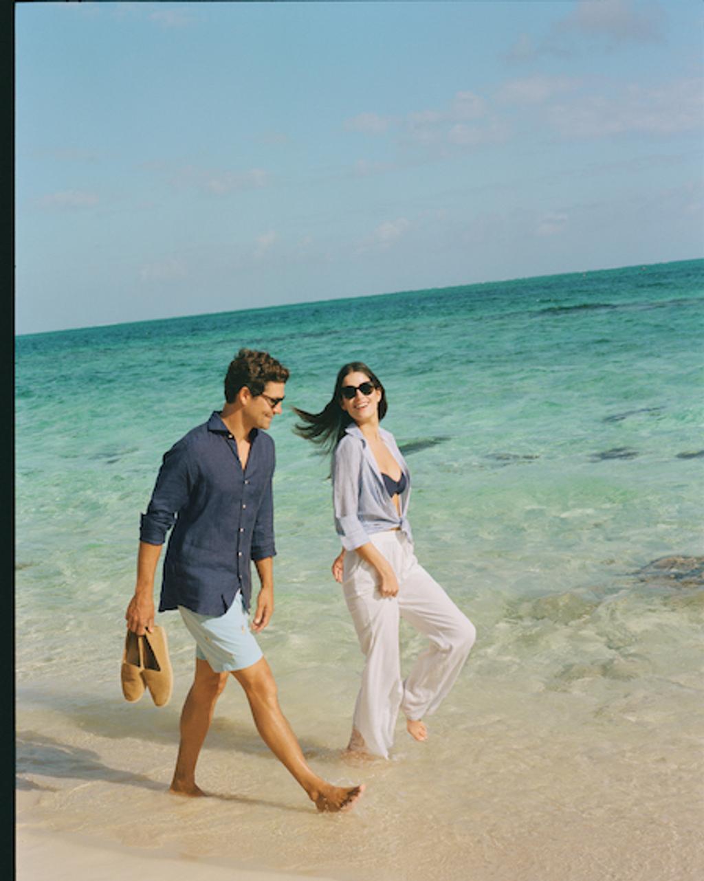 Man in navy shirt and blue shorts and woman in blue shirt and white pants walking on the beach