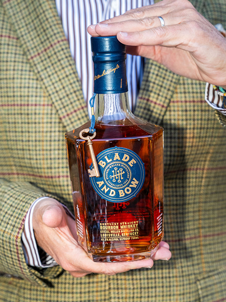 Man in patterned suit holding a bottle of "Blade and Bow" whiskey.