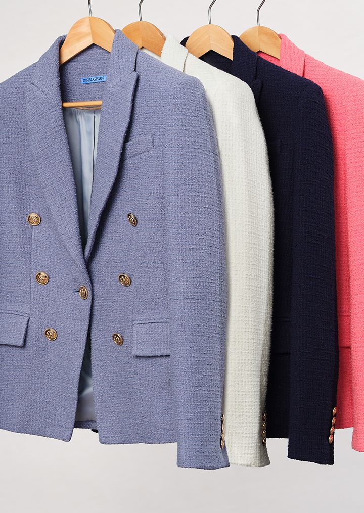 Four double-breasted tweed blazers with gold buttons in light blue, white, navy, and pink, hanging on hangers.