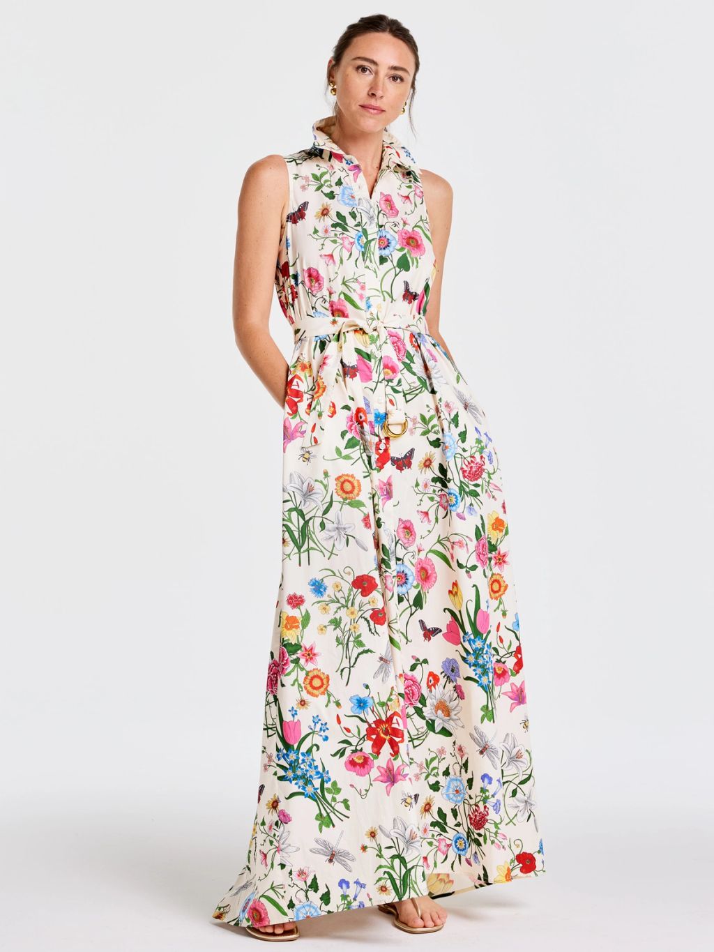 Woman in a sleeveless, collared, off-white maxi dress with a colorful floral and insect pattern and a belted waist.
