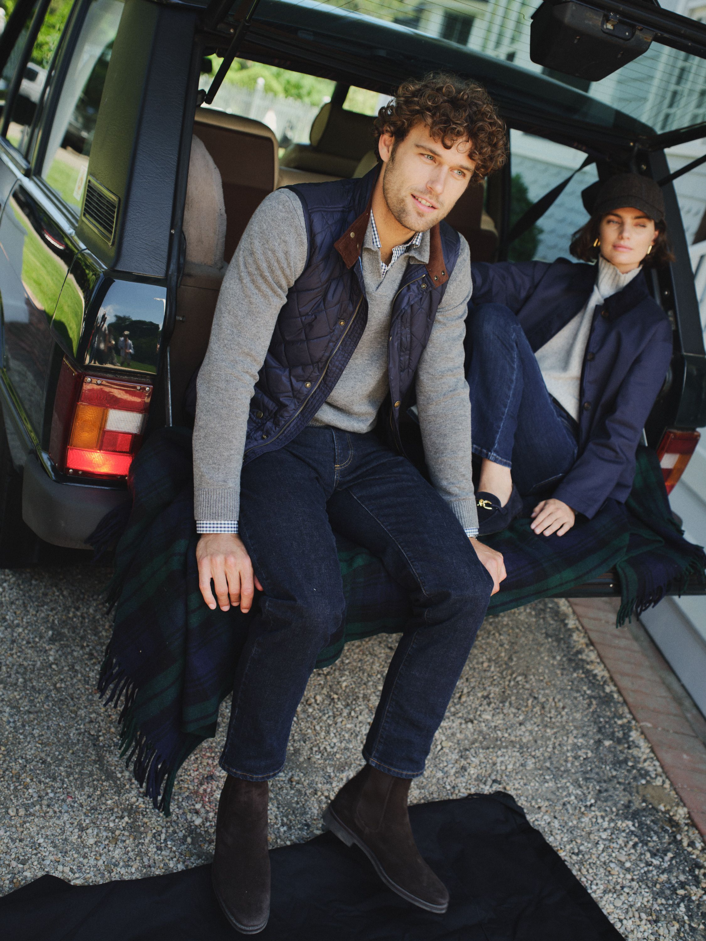 A man and woman sit on the open tailgate of a dark SUV, casually dressed in layered clothing, with a plaid blanket.
