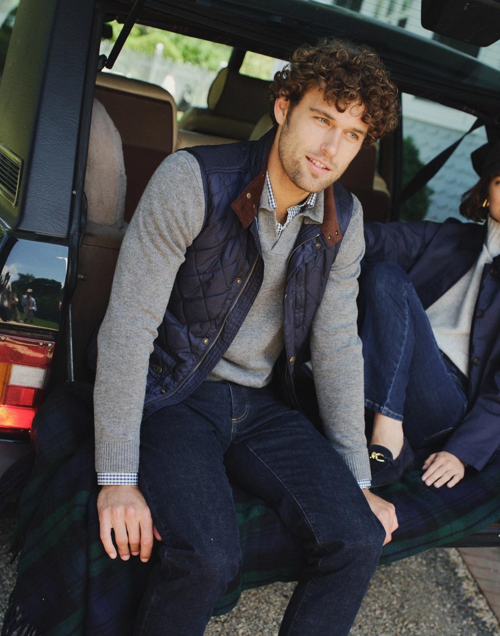 A man and woman sit on the open tailgate of a dark SUV, casually dressed in layered clothing, with a plaid blanket.