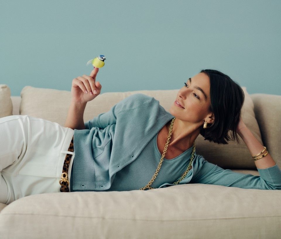 A woman lies on a beige sofa, smiling at a small blue and yellow bird perched on her extended finger.