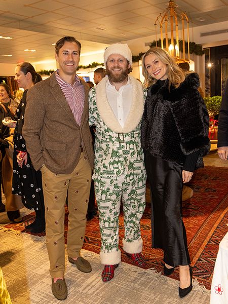 Three people in formal attire pose together at an upscale holiday event or party. Holiday decorations including garland are visible in the background of what looks like a retail or boutique setting.
