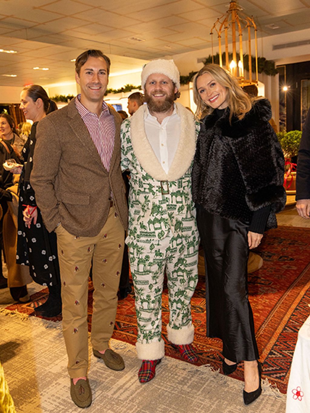 Three people in formal attire pose together at an upscale holiday event or party. Holiday decorations including garland are visible in the background of what looks like a retail or boutique setting.