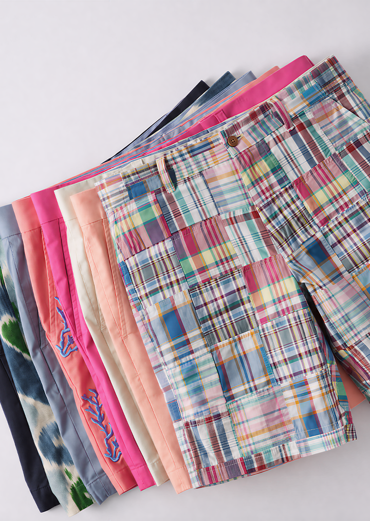 Seven colorful pairs of shorts, including solid colors, tie-dye, and patchwork plaid patterns, stacked diagonally on a white background.