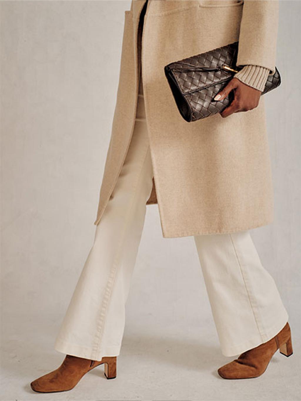 Women with brown heeled boots, white pants, and a cream colored trench coat walking while holding a brown leather clutch.