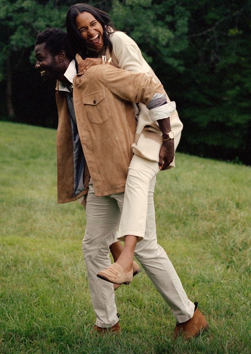 man and woman walking through field, him carrying her on his back. He is wearing tan suede safari style jacket with khaki chino pants