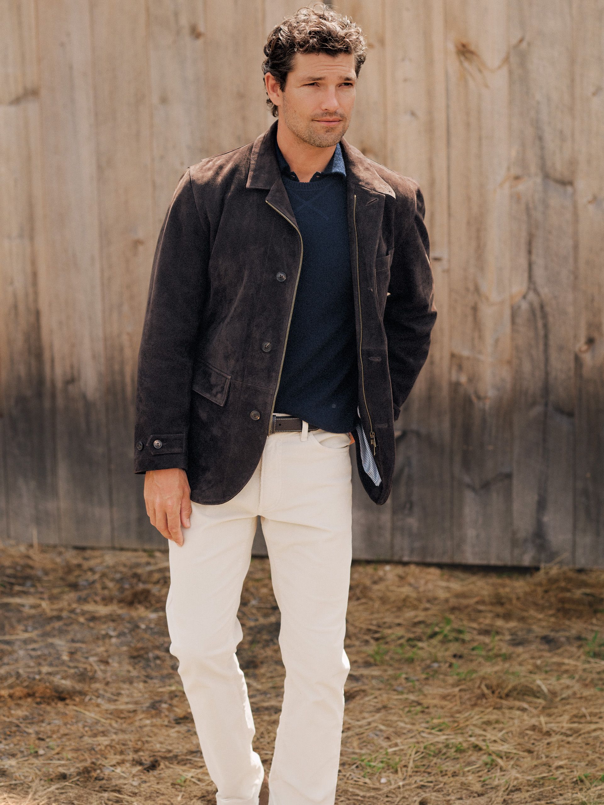 A man in a dark brown suede jacket, navy sweater, and cream pants stands in front of a wooden fence.