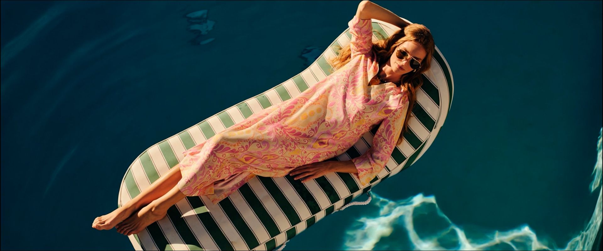 A woman in a pink dress and sunglasses floats on a striped raft in a swimming pool.