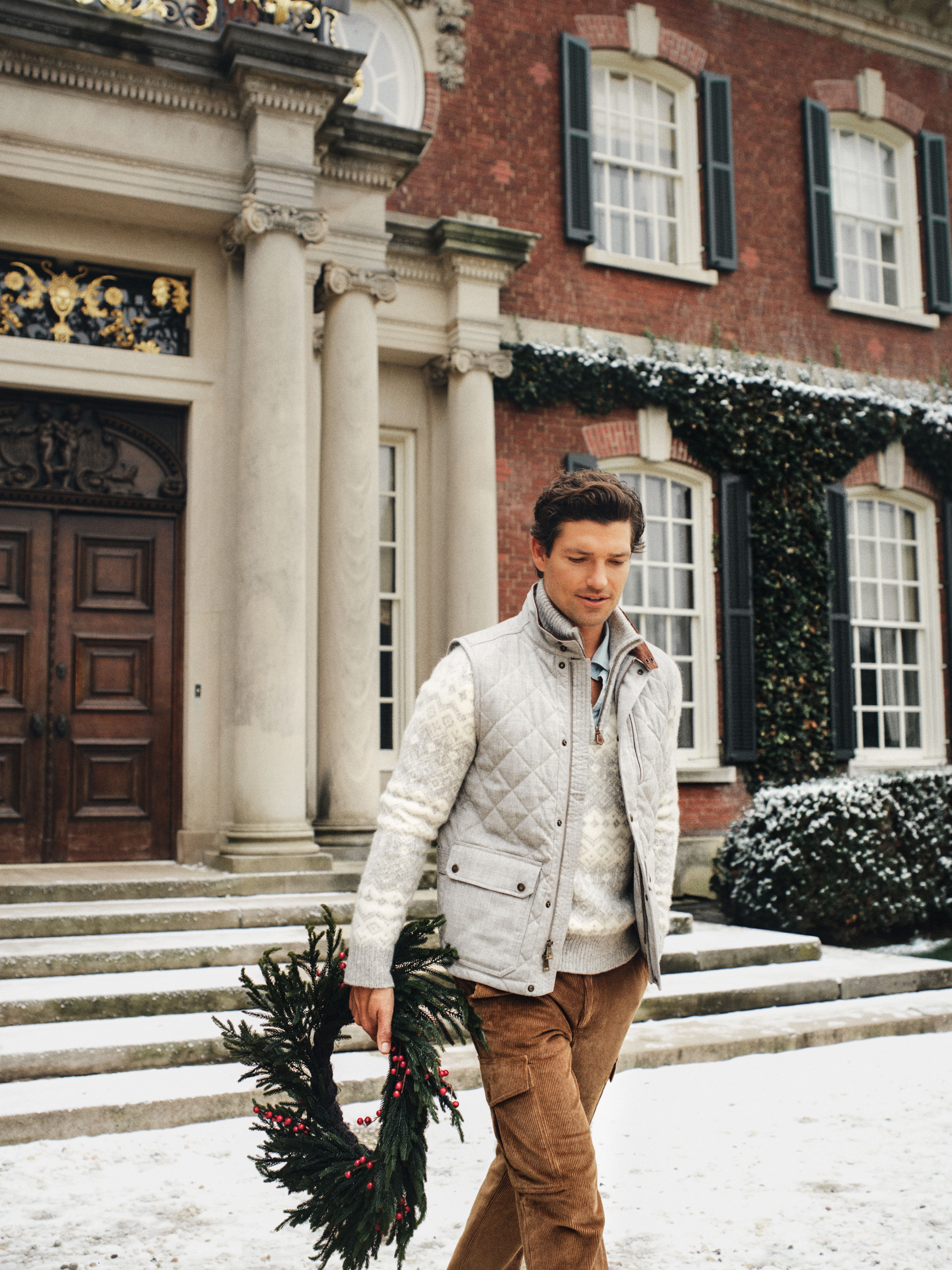 A man in a quilted vest and sweater walks through snow in front of a grand brick house, holding a holiday wreath.