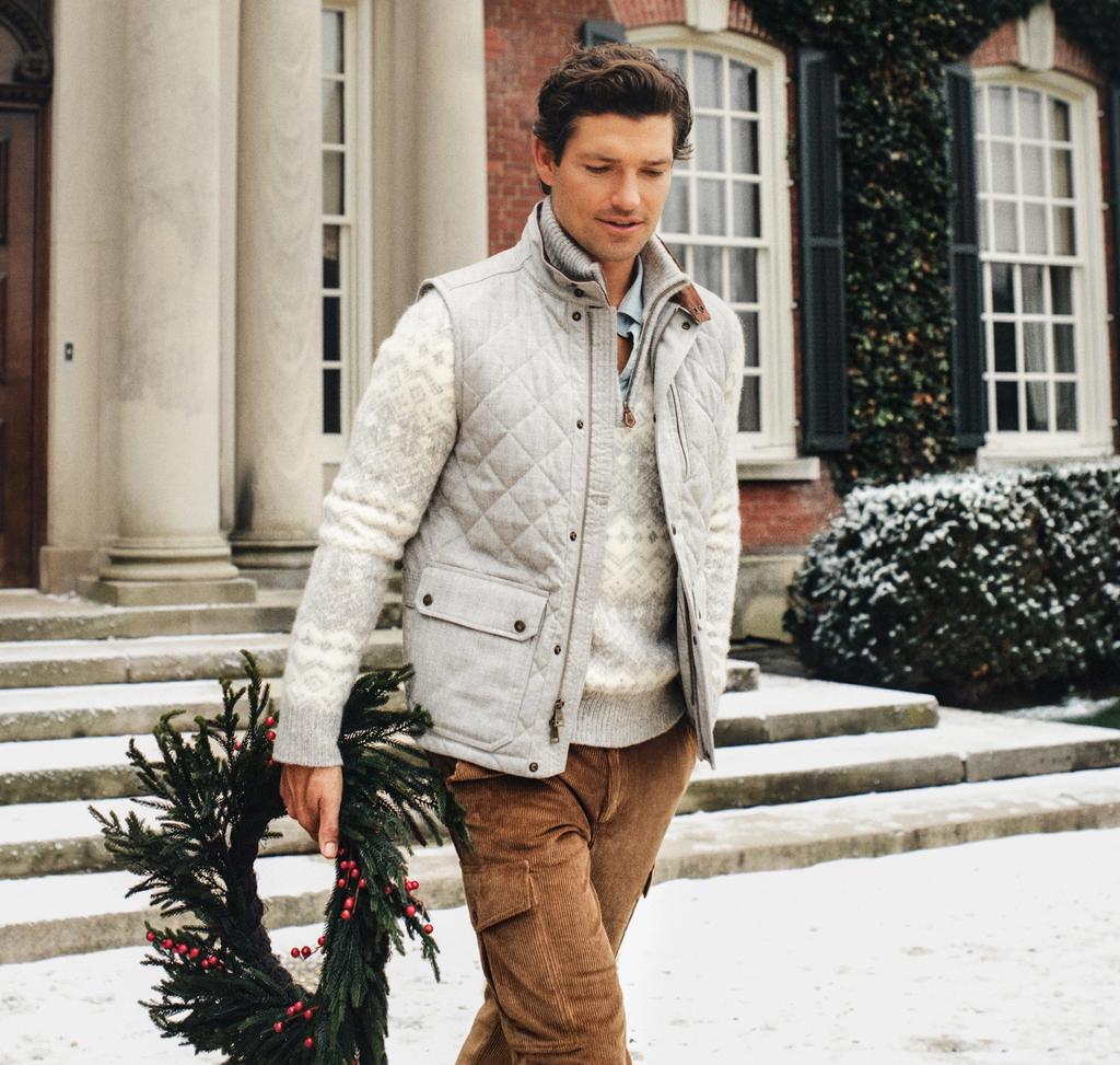 A man in a quilted vest and sweater walks through snow in front of a grand brick house, holding a holiday wreath.