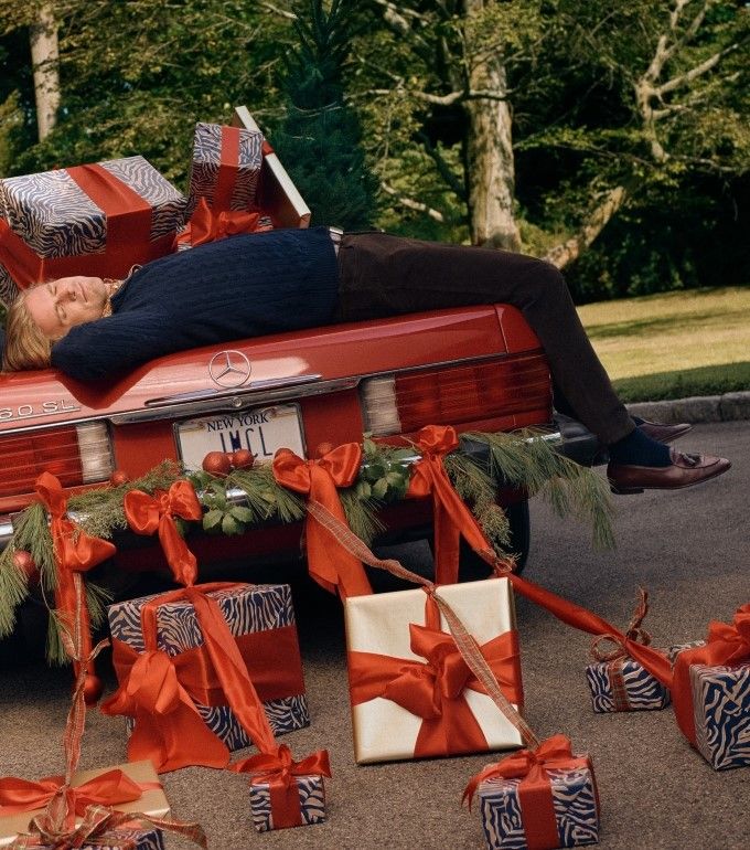 man laying on red convertible car sleeping and surrounded by wrapped holiday gifts