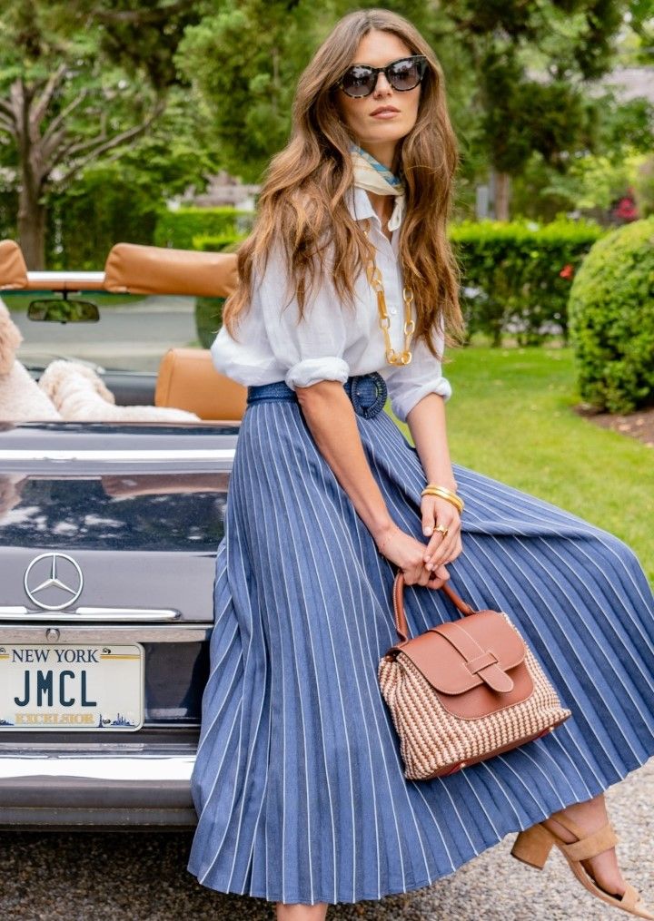 woman seated on back of vintage looking car, outside, wearing navy pleated skirt and white linen shirt