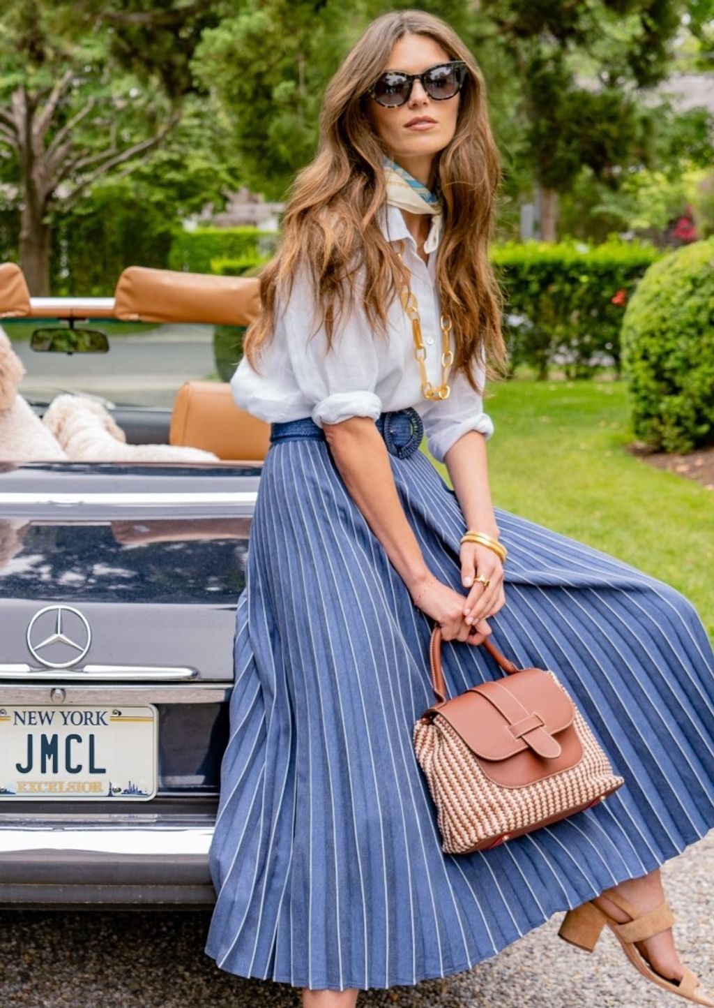woman seated on back of vintage looking car, outside, wearing navy pleated skirt and white linen shirt